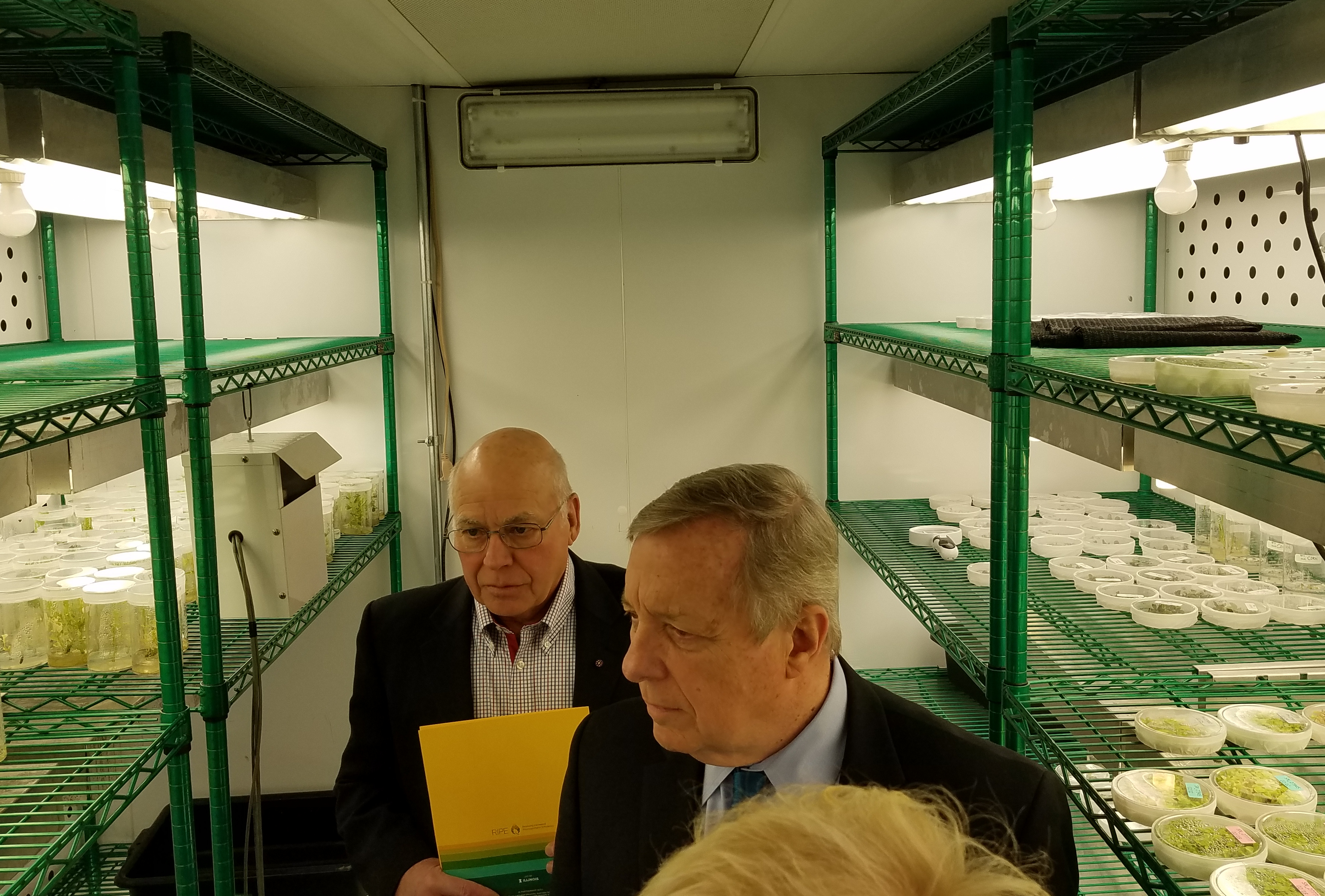 Don Ort and Dick Durbin are inside a growth chamber with shelves holding petri dishes containing small plants.