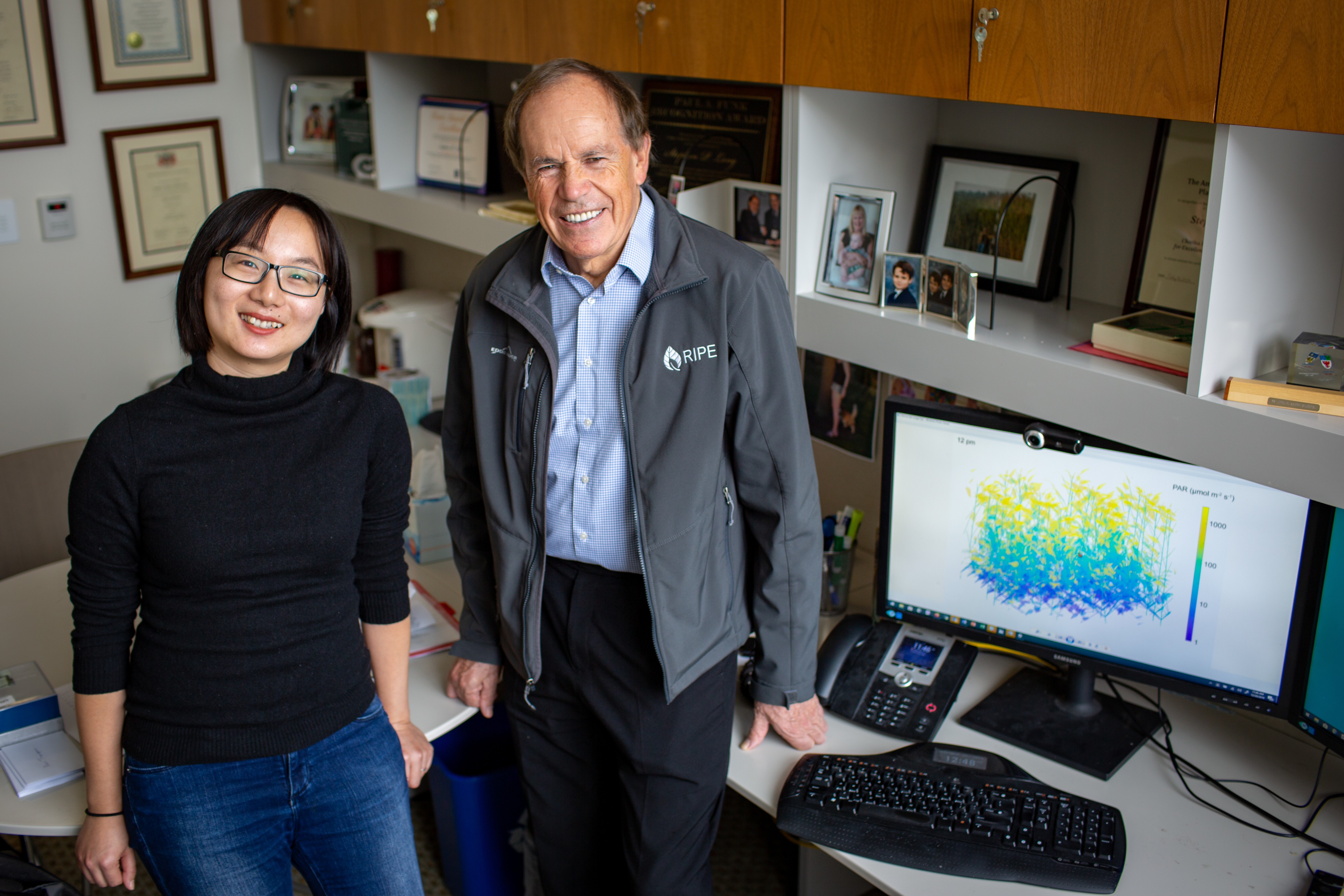 Yu Wang and Steve Long sit on edge of desk next to a computer screen displaying their model. 